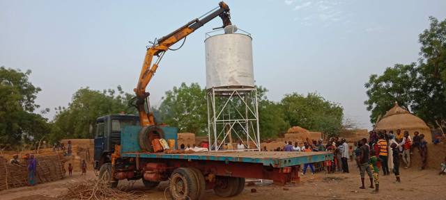Une grue pose un réservoir sur sa tour métallique, devant un village